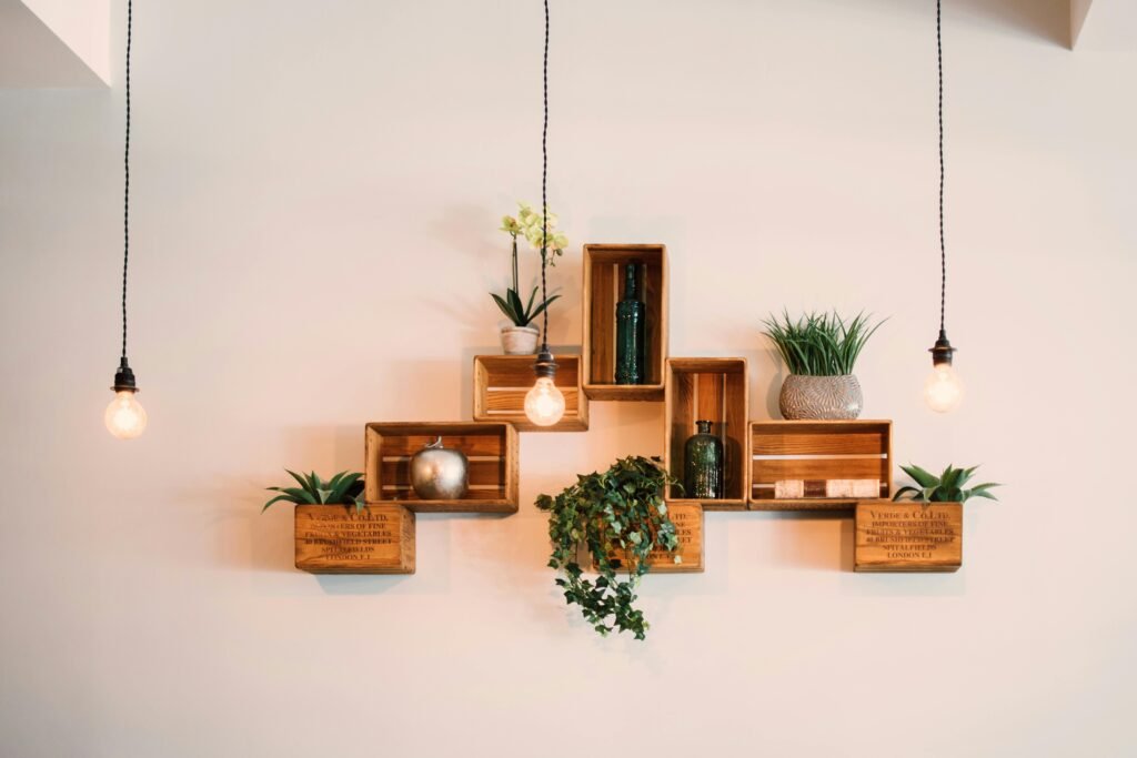 A decorative wall display featuring rustic wooden crates transformed into shelves, holding various plants, glass bottles, and a silver pumpkin, flanked by hanging Edison-style pendant lights on black cords